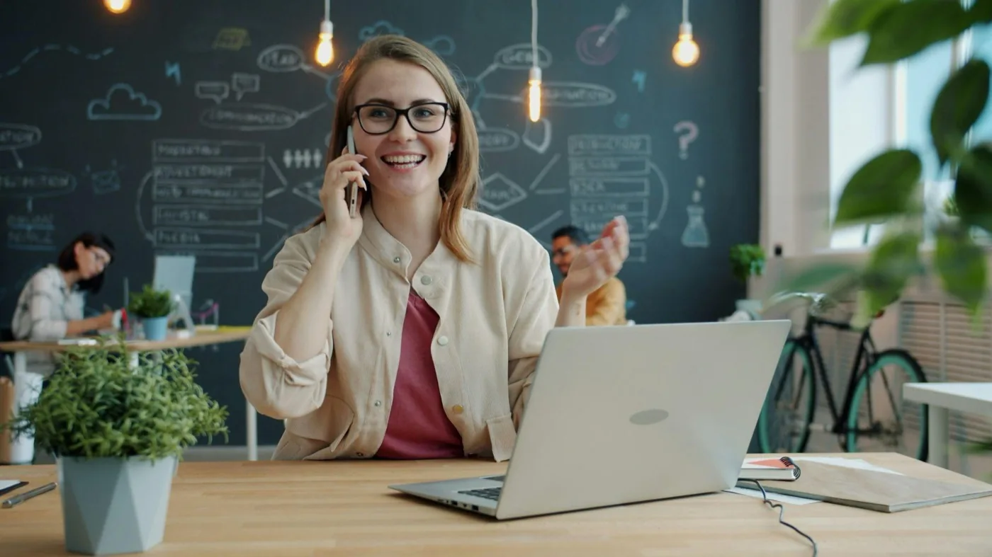 A smiling woman on a phone call at her desk, gesturing, with a laptop open in front of her