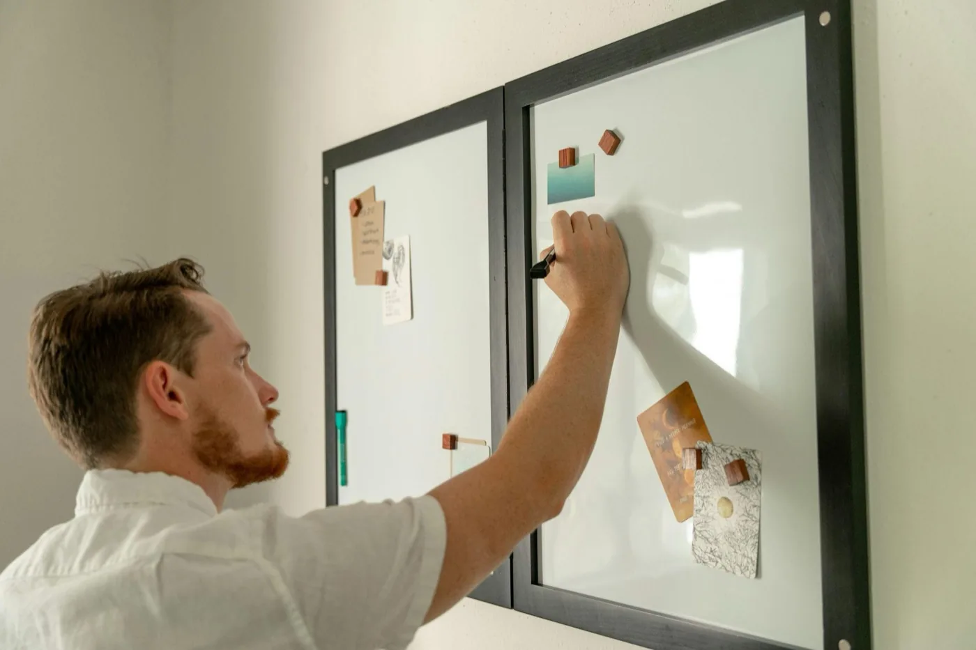 A man writing on a glass whiteboard covered in pinned notes and cards