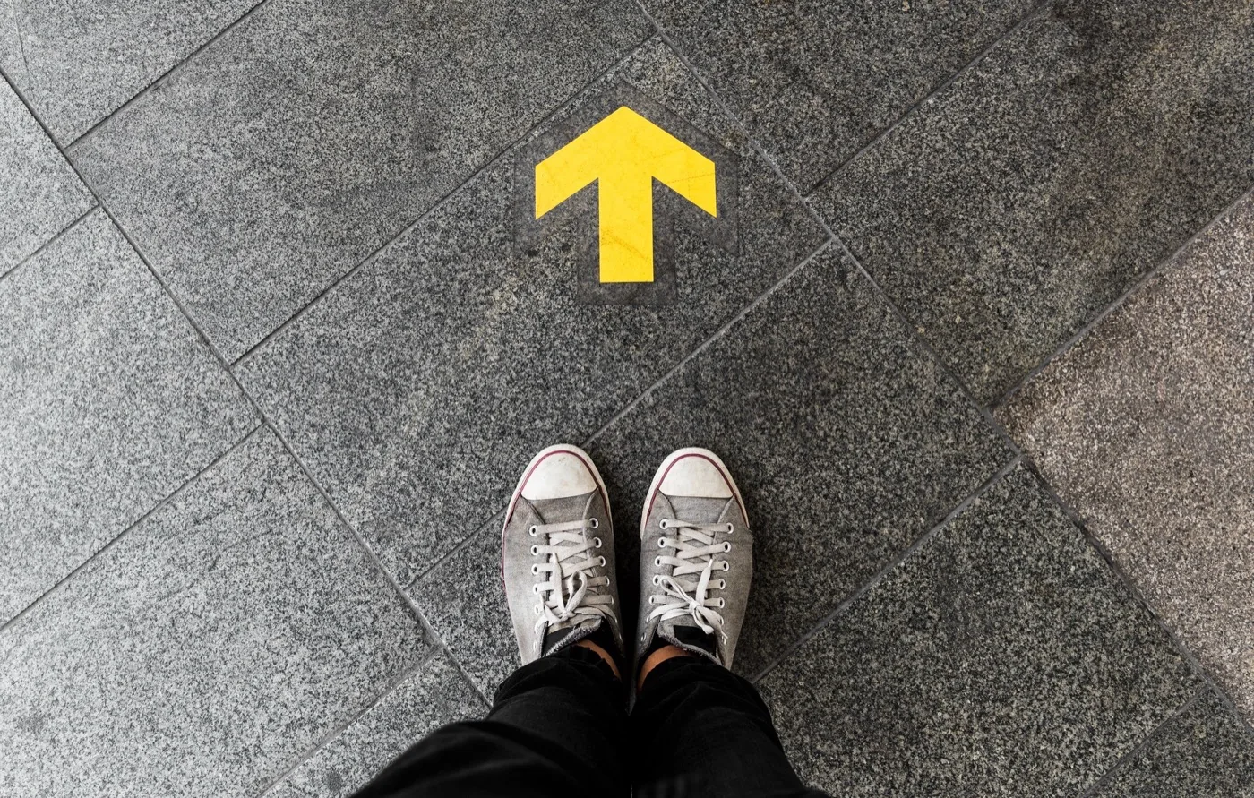 Looking down at a pair of feet standing on a yellow arrow painted on pavement