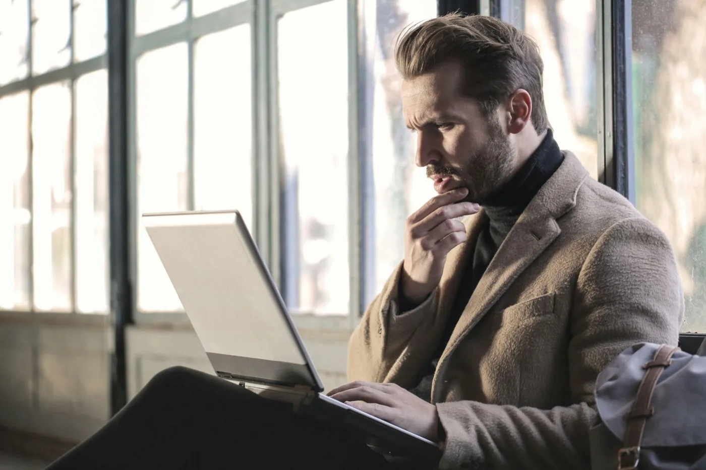 A man in a camel coat frowning at his laptop with his hand on his chin, deep in thought