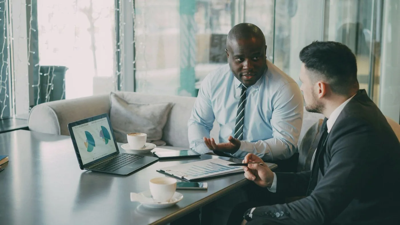Two businessmen in conversation over coffee, reviewing charts on a laptop and printed reports