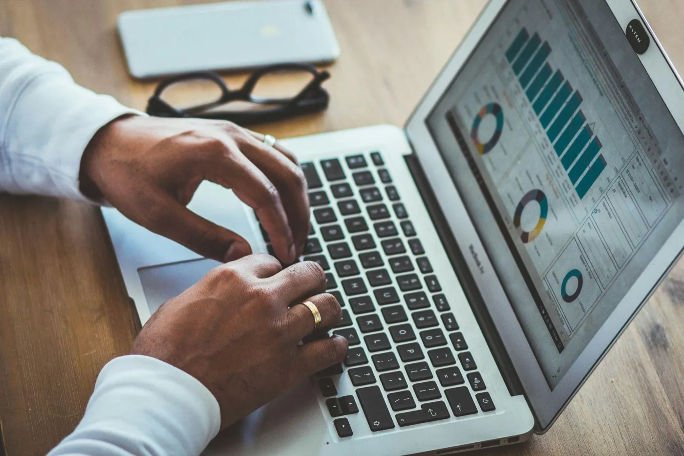 Hands typing on a laptop showing bar and doughnut charts, with glasses and a phone nearby