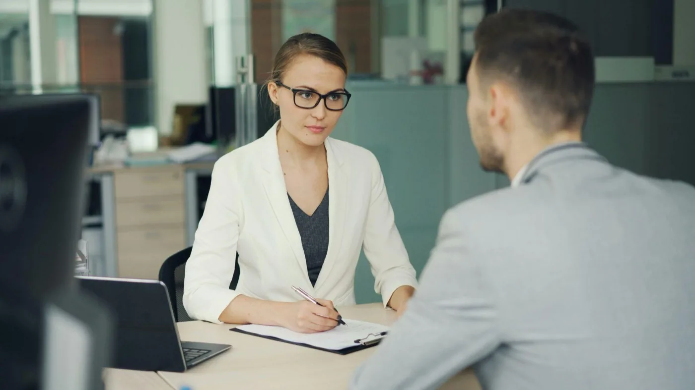 A woman in glasses taking notes on a clipboard while interviewing a man across a desk