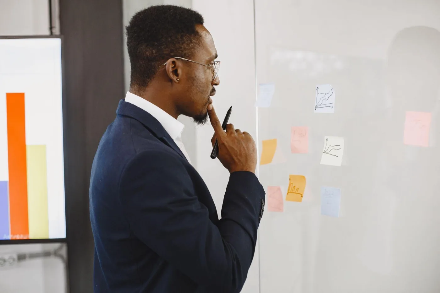 A man in a blazer studying charts and sticky notes on a glass whiteboard, pen at his chin