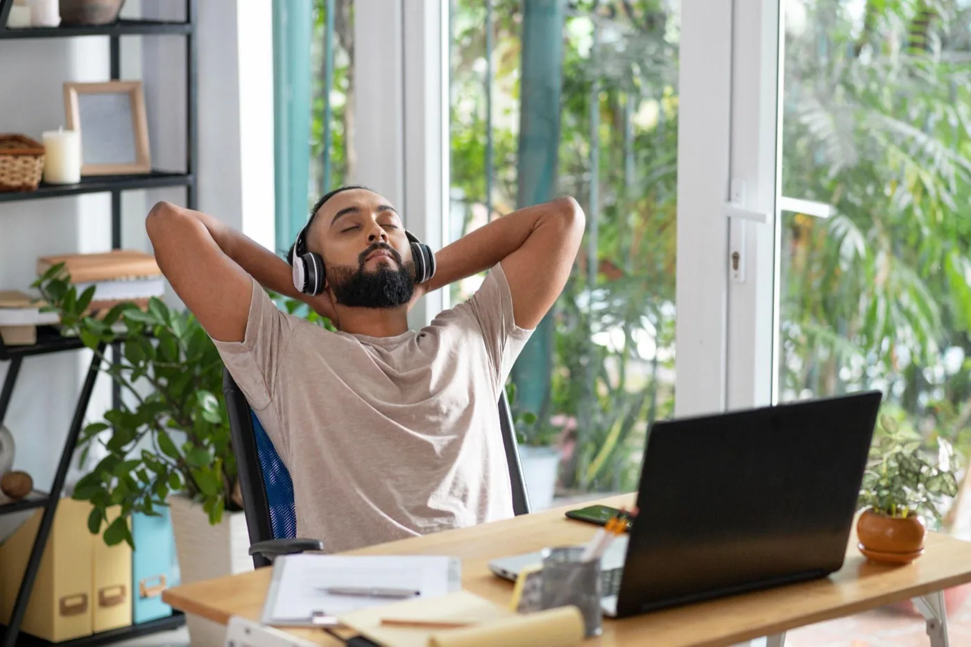 A relaxed man leaning back at his desk with headphones on and a laptop open in front of him