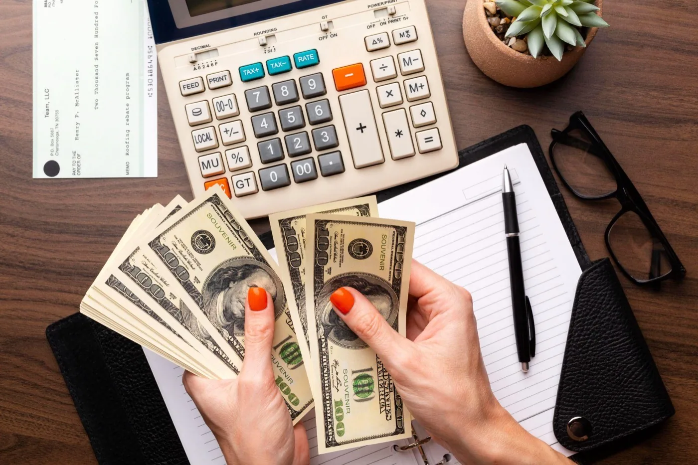 A person counting hundred-dollar bills over a calculator, notebook and cheque on a desk